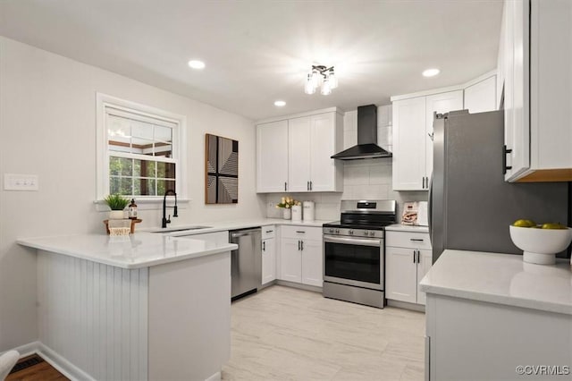 kitchen with a peninsula, wall chimney range hood, white cabinets, a sink, and stainless steel appliances