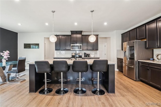 kitchen featuring tasteful backsplash, a kitchen island with sink, stainless steel appliances, light wood-style flooring, and light stone countertops