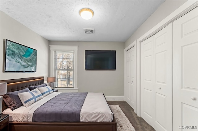 bedroom with baseboards, multiple closets, a textured ceiling, dark carpet, and visible vents