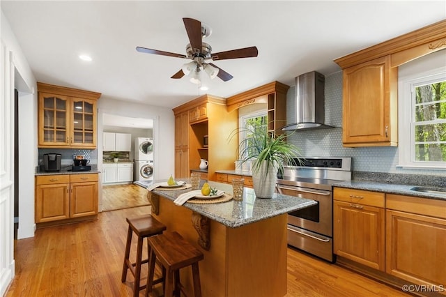 kitchen featuring stacked washer and clothes dryer, backsplash, a ceiling fan, wall chimney exhaust hood, and stainless steel electric range