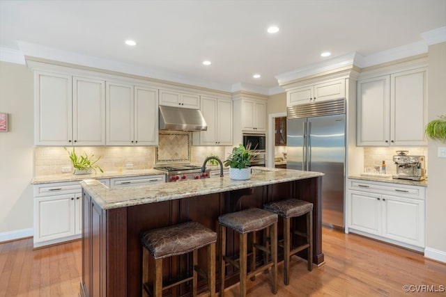 kitchen featuring decorative backsplash, under cabinet range hood, built in appliances, and a kitchen breakfast bar