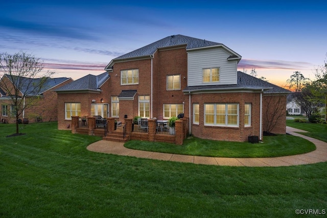back of house at dusk with a yard, a wooden deck, brick siding, and roof with shingles