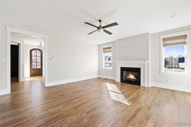unfurnished living room featuring a warm lit fireplace, a wealth of natural light, a ceiling fan, and light wood finished floors