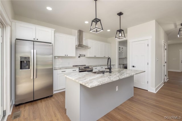 kitchen featuring wood finished floors, a sink, a center island with sink, wall chimney exhaust hood, and appliances with stainless steel finishes