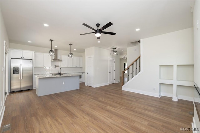 kitchen with wall chimney range hood, a kitchen island with sink, ceiling fan, stainless steel refrigerator with ice dispenser, and recessed lighting