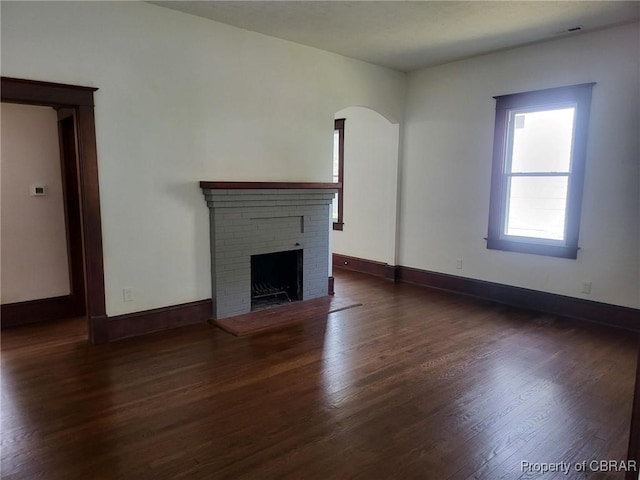 unfurnished living room featuring arched walkways, wood finished floors, baseboards, and a brick fireplace
