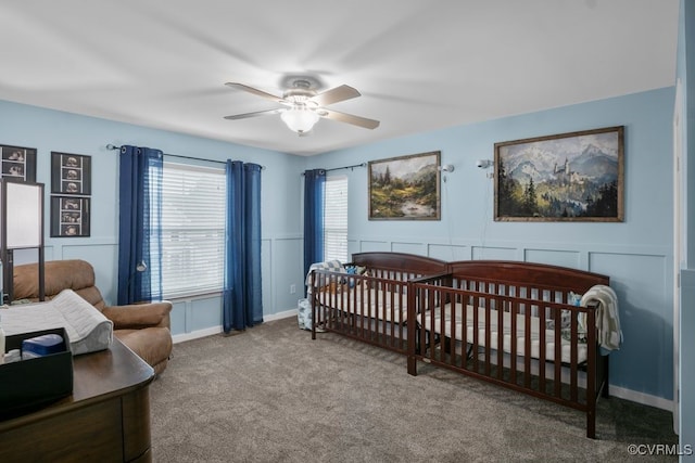 bedroom featuring a ceiling fan, a wainscoted wall, a crib, a decorative wall, and carpet flooring