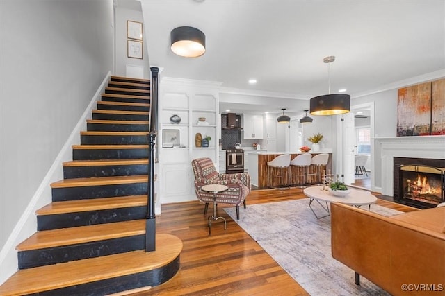 living area featuring stairway, wood finished floors, a warm lit fireplace, ornamental molding, and recessed lighting