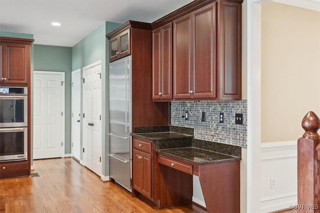 kitchen featuring appliances with stainless steel finishes, dark stone counters, light wood-type flooring, backsplash, and glass insert cabinets