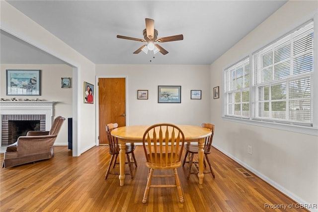 dining room featuring wood finished floors, ceiling fan, baseboards, and a brick fireplace