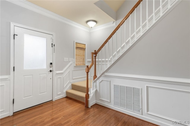 entrance foyer featuring crown molding, a decorative wall, stairs, light wood-type flooring, and wainscoting