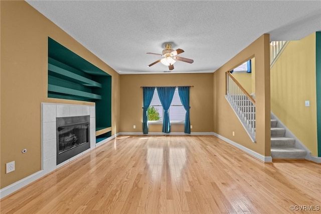 unfurnished living room featuring a tiled fireplace, a ceiling fan, a textured ceiling, wood finished floors, and stairs