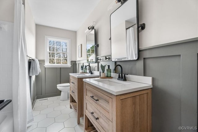 bathroom featuring toilet, a shower with shower curtain, two vanities, a wainscoted wall, and a sink
