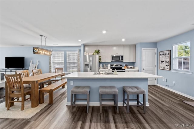 kitchen with appliances with stainless steel finishes, a center island with sink, a sink, light countertops, and dark wood-type flooring
