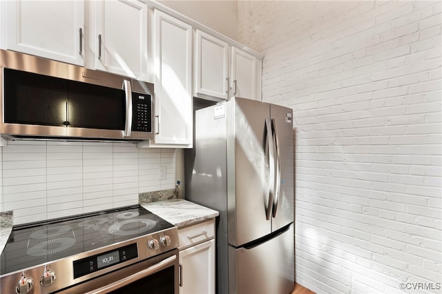 kitchen featuring appliances with stainless steel finishes, light stone counters, brick wall, white cabinetry, and tasteful backsplash