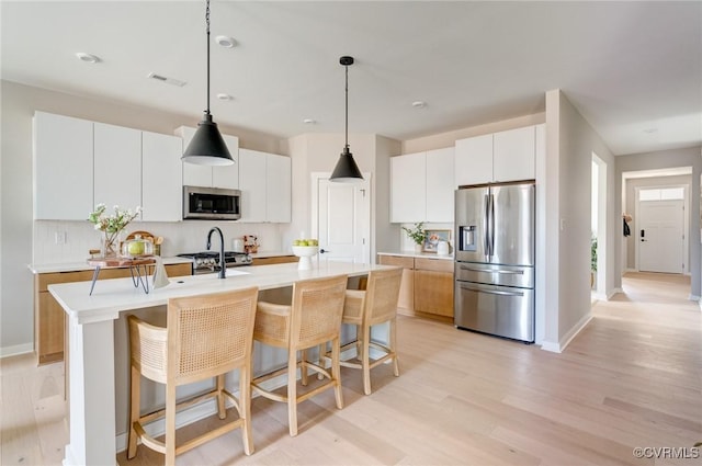kitchen featuring appliances with stainless steel finishes, light countertops, an island with sink, decorative backsplash, and light wood-type flooring