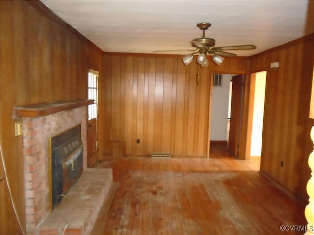 unfurnished living room featuring a ceiling fan, visible vents, wood finished floors, a brick fireplace, and wooden walls