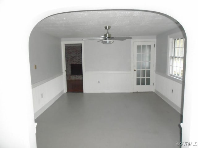 empty room featuring a ceiling fan, a textured ceiling, arched walkways, a wainscoted wall, and a fireplace