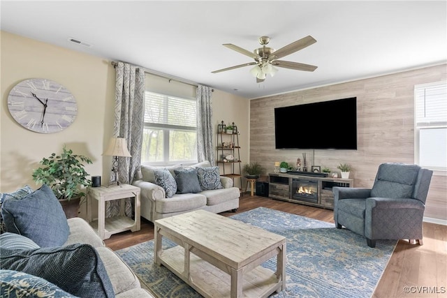 living area with ceiling fan, an accent wall, wood finished floors, and a glass covered fireplace