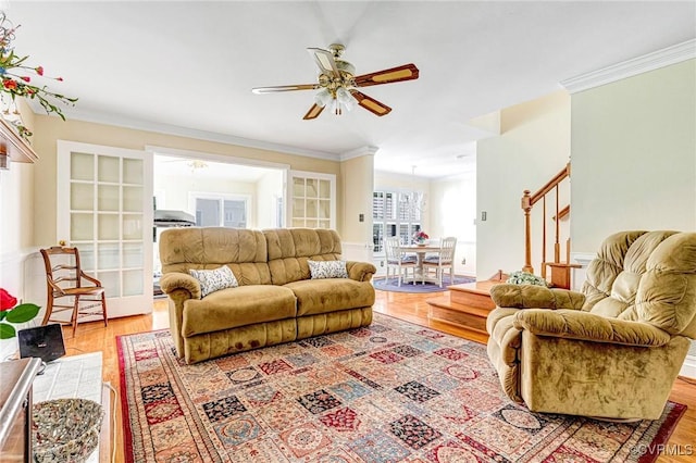 living area featuring crown molding, light wood-style flooring, ceiling fan, and stairway