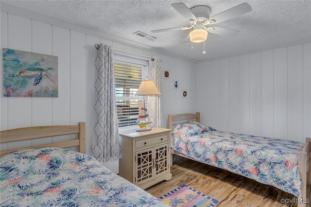 bedroom with a textured ceiling, visible vents, a ceiling fan, wood finished floors, and a decorative wall