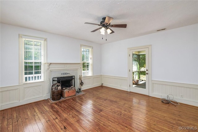 unfurnished living room featuring ceiling fan, visible vents, a fireplace, hardwood / wood-style flooring, and a wainscoted wall