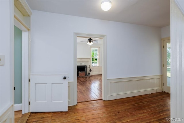 empty room featuring wainscoting, a fireplace with raised hearth, a ceiling fan, and hardwood / wood-style floors