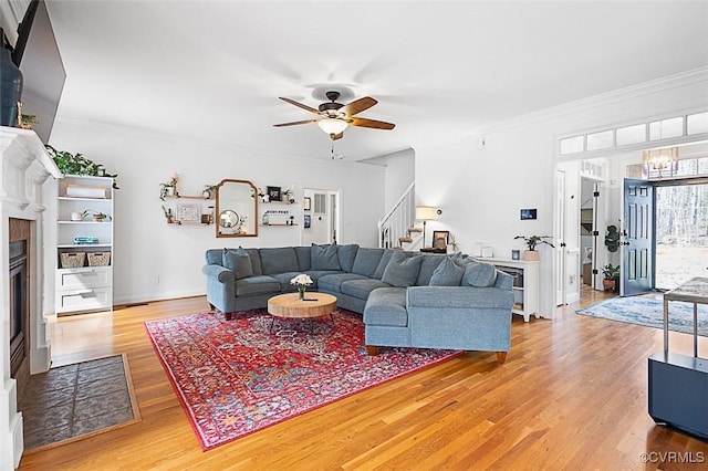 living room featuring a tiled fireplace, light wood finished floors, stairway, a ceiling fan, and crown molding