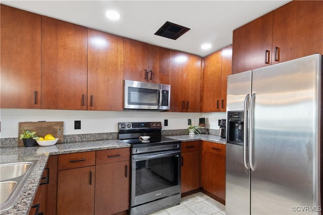 kitchen with a sink, brown cabinets, light tile patterned flooring, stone countertops, and appliances with stainless steel finishes