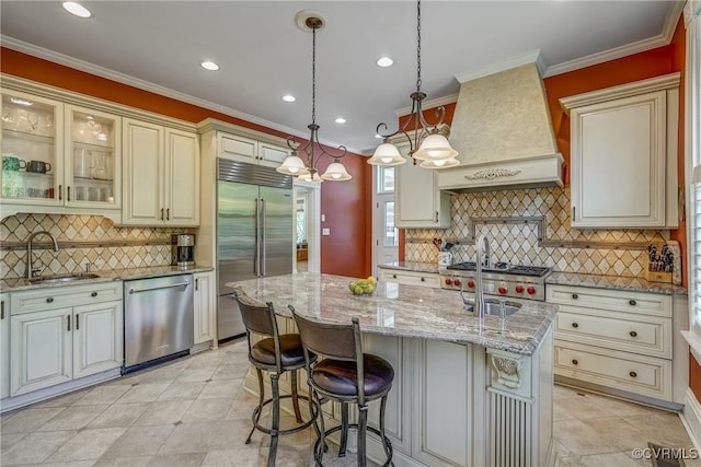 kitchen with decorative backsplash, premium range hood, cream cabinetry, a sink, and stainless steel appliances