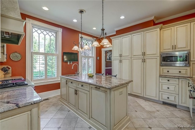 kitchen featuring a center island with sink, light tile patterned floors, stainless steel microwave, ornamental molding, and cream cabinetry