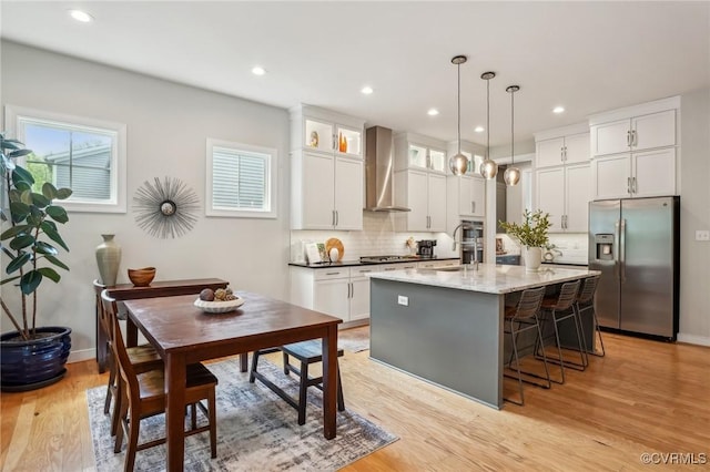 kitchen with wall chimney range hood, tasteful backsplash, light wood-type flooring, a center island with sink, and appliances with stainless steel finishes