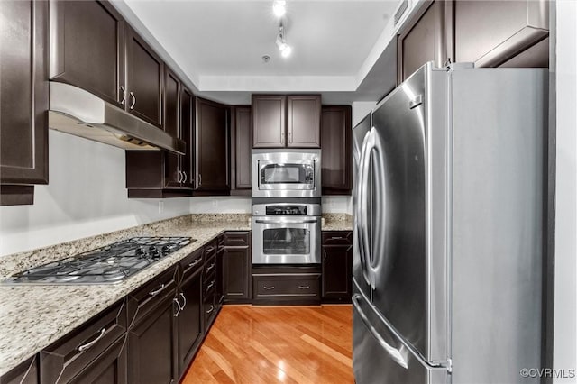 kitchen with light stone counters, under cabinet range hood, stainless steel appliances, dark brown cabinets, and light wood-style flooring