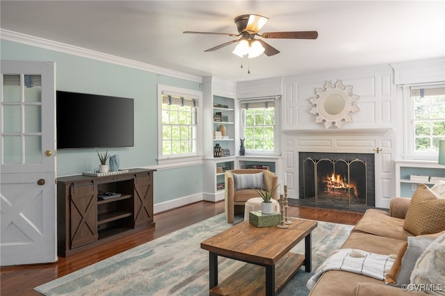 living area featuring a warm lit fireplace, a ceiling fan, baseboards, wood finished floors, and ornamental molding