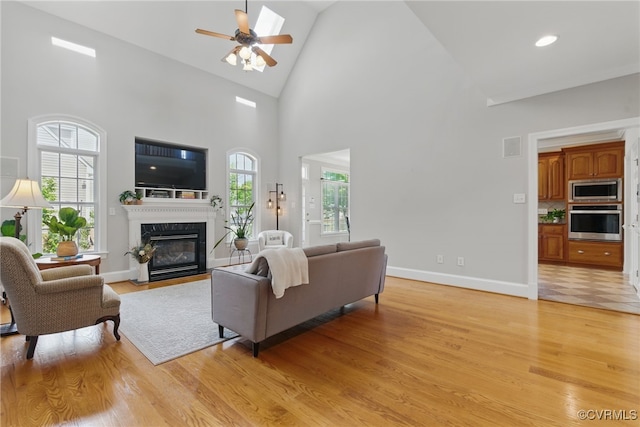 living room featuring light wood finished floors, baseboards, high vaulted ceiling, a ceiling fan, and a fireplace