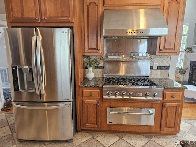 kitchen with a warming drawer, stainless steel refrigerator with ice dispenser, dark stone countertops, and exhaust hood
