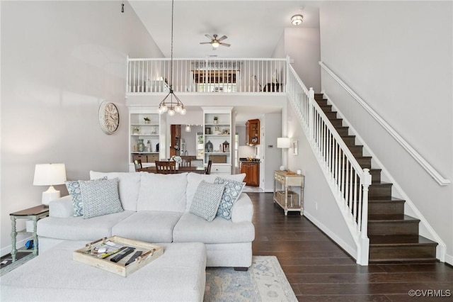 living room with ceiling fan with notable chandelier, stairs, baseboards, a towering ceiling, and dark wood-type flooring