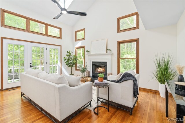living room featuring high vaulted ceiling, a warm lit fireplace, a ceiling fan, light wood-style flooring, and baseboards