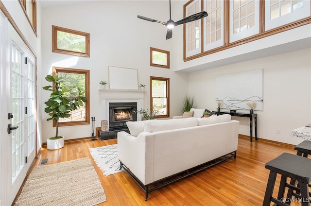 living area with a high ceiling, a lit fireplace, light wood finished floors, baseboards, and a ceiling fan