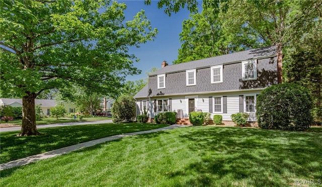 colonial inspired home with a gambrel roof, a front yard, a chimney, and a shingled roof