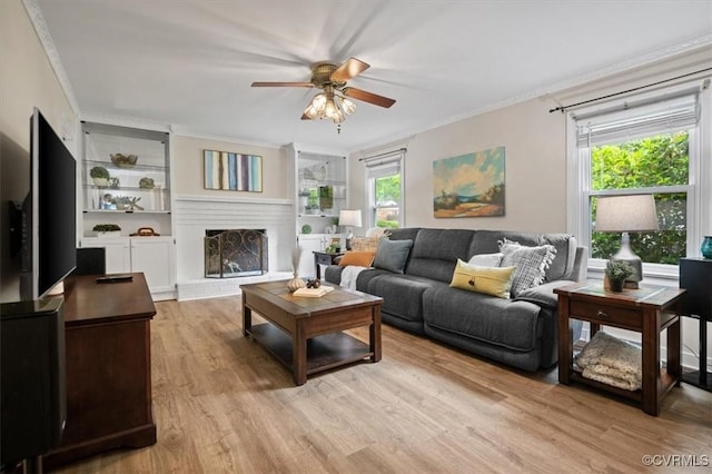 living area featuring crown molding, a ceiling fan, a brick fireplace, light wood-style floors, and built in shelves