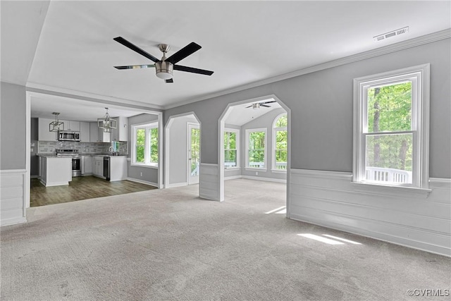 unfurnished living room featuring visible vents, a wainscoted wall, ceiling fan with notable chandelier, ornamental molding, and dark carpet