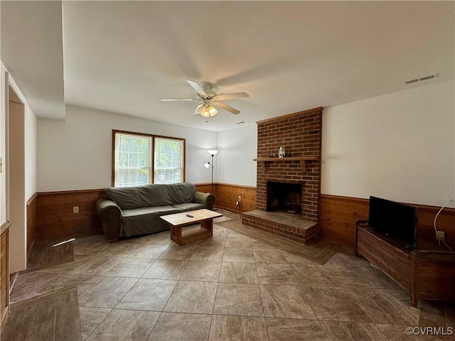 living room with wooden walls, wainscoting, a fireplace, a ceiling fan, and visible vents