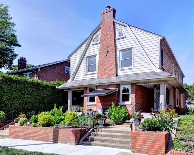 view of front of property featuring a porch, a gambrel roof, a chimney, a ceiling fan, and brick siding