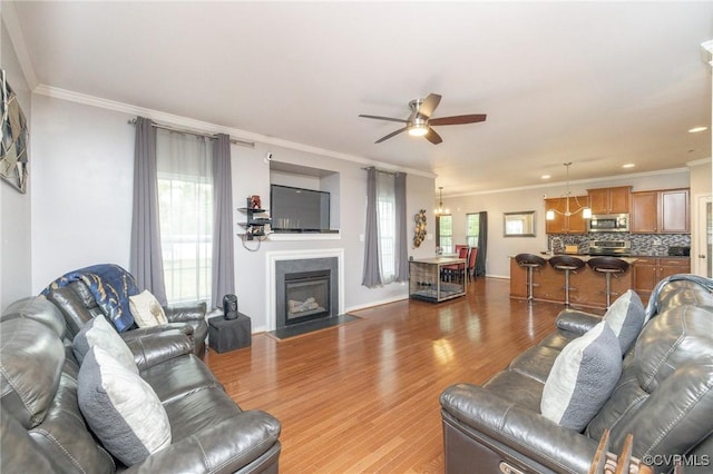 living area featuring a fireplace with flush hearth, wood finished floors, ornamental molding, and ceiling fan with notable chandelier