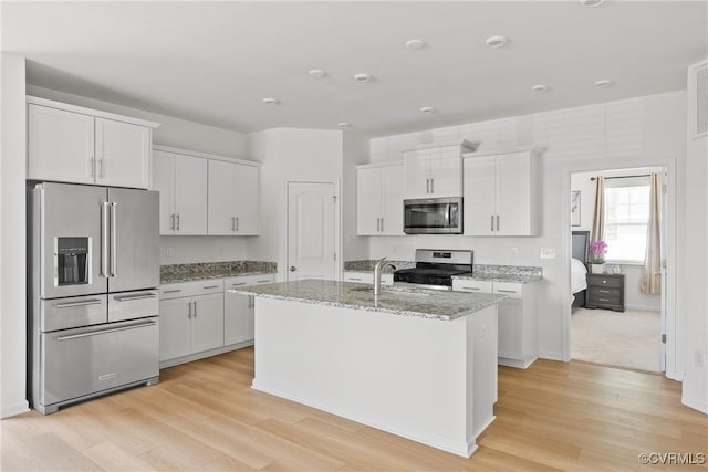kitchen featuring light stone counters, an island with sink, light wood-style flooring, stainless steel appliances, and white cabinetry