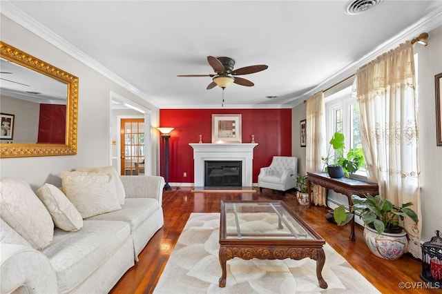 living area featuring wood finished floors, a glass covered fireplace, visible vents, a ceiling fan, and crown molding