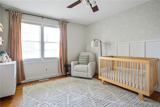 bedroom with wainscoting, wood finished floors, ceiling fan, wallpapered walls, and a crib
