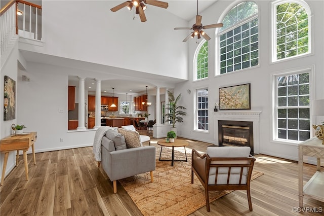 living room with a high ceiling, ceiling fan, a glass covered fireplace, light wood-style flooring, and ornate columns