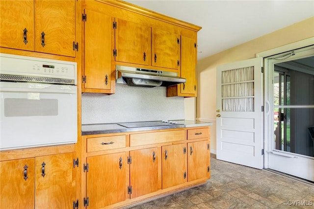 kitchen featuring white oven, black electric cooktop, under cabinet range hood, brown cabinets, and stone finish flooring